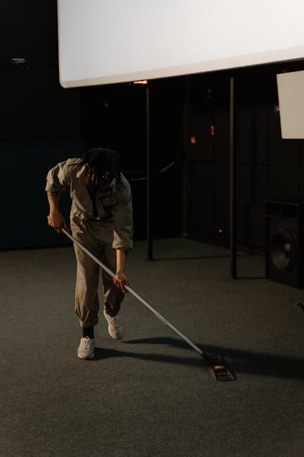 A person dressed in a grey uniform is performing surface cleaning on a carpeted floor using a long-handled tool in a room with dark walls and minimal lighting. The scene appears to be in a commercial 
