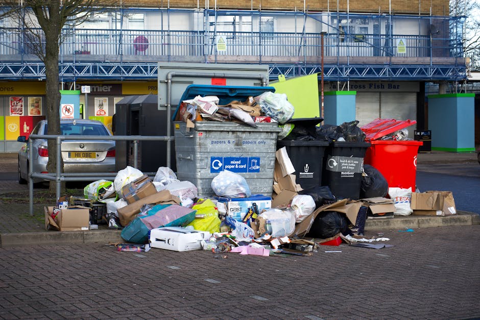Outdoor scene showing a large metal recycling bin labeled 'mixed paper & card' overflowing with discarded paper, cardboard boxes, plastic bags, and packaging materials. Surrounding the bin are additio