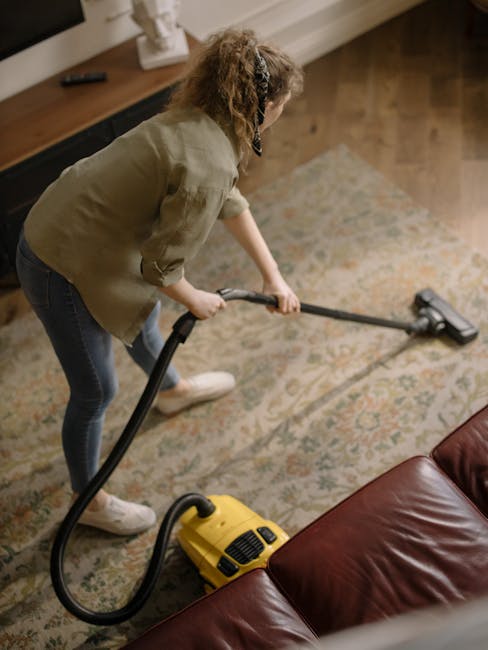 A woman using a yellow vacuum cleaner with a hose attachment to deep clean a patterned carpet in a living room. She is wearing a light-colored jacket, dark jeans, and white shoes, and is focused on su